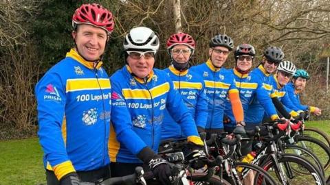 The cyclists are lined up with their bikes and wear bright blue and yellow cycling tops with 100 laps for Rob on them. They wear helmets of varying colours.