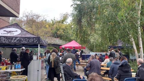 A crowd of people at Mount Place during Jericho's Oktoberfest on 17 October. Some are buying food and drinks while others are watching a band perform. A tent branded Little Ox Brew can be seen. It is cloudy.