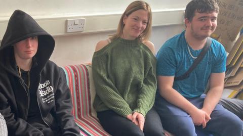 Four people sitting in a line on a striped sofa - the woman on the left has blonde hair and a grey top. The woman next to her is dressed all in black with a hood up. The next woman is blonde and wearing a green top and black trousers. The man on the right has a blue shite, a necklace, blue trousers and blue trainers.