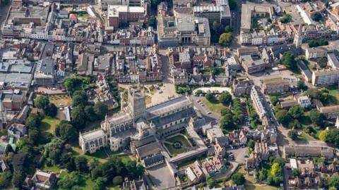 An aerial shot of Gloucester city centre on a sunny day, including Gloucester Cathedral in the foreground, green spaces, Georgian buildings, and Shire Hall
