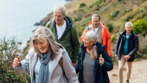 A group of people walk along the Cornish coast. There are two women and three men total. All are wearing wet-weather gear and are smiling.