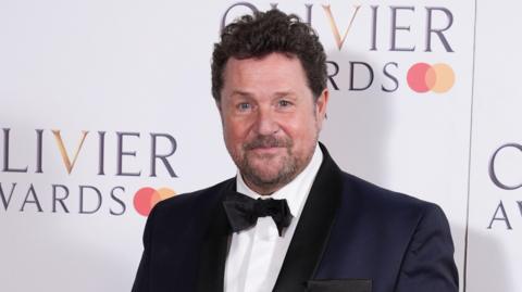 Michael Ball in a black tuxedo. He is attending the Olivier Awards and is standing in front of a white board which says OLIVIER AWARDS. 