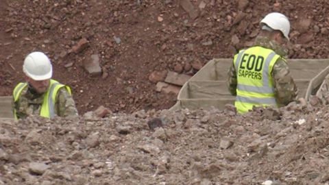 Two people in Army fatigues and high-vis jackets with white hat in a hole