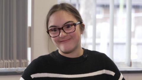 Laura has dark hair and dark rimmed glasses and is wearing a black and white striped jumper