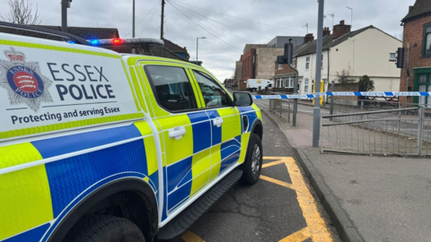 An Essex Police car has been parked on a road in front of blue and white police tape. A section of the road has been cordoned off. 