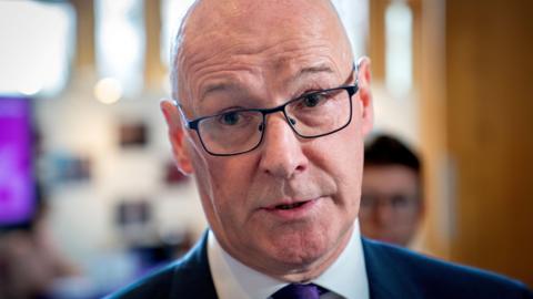 John Swinney - a bald man with glasses and wearing a suit - speaks to media in a lobby