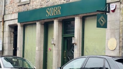 The front of a bar called Sobr, gold lettering on a green background, and cars are parked in front.