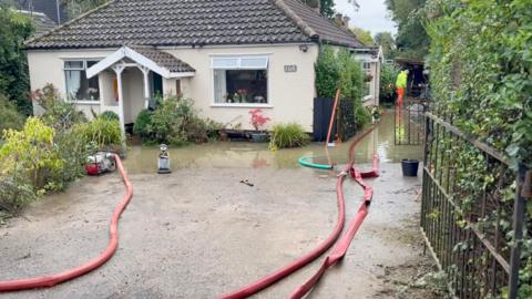 A bungalow is surrounded by several feet of water. Three fire hoses trail along the ground to pump out the water.