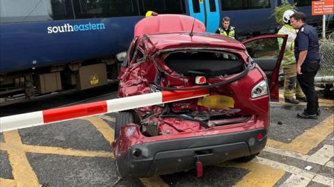 A red damaged car behind a closed red and white bar. A navy train can be seen behind it.