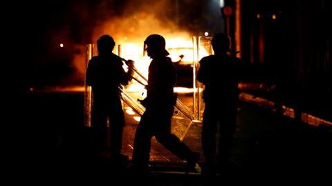 The silhouettes of three riot police standing in front of a burning vehicle 