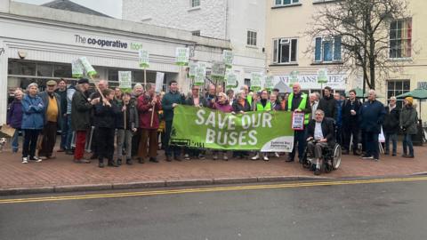 A crowd of about 50 people gathered together holding placards and a banner reading 'save our buses'