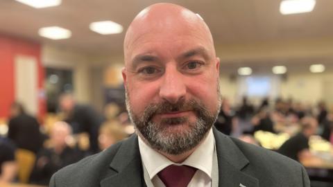 Head and shoulders shot of Marc Jones. He is wearing a grey suit and red tie and is looking directly at the camera. The background of the image is blurred but shows people sitting and standing at desks.