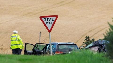 A police officer stands at a junction in the road between green grass and a brown crop field. Next to him there is a give way sign, a black Volkswagen Golf and the wreckage of another car. 