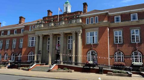 A large three-storey red brick council house building with large double-storey height pillars flanking the main door and Victorian lanterns lining ramps to the left and right. It is a sunny day and a person in shorts sits outside. Bunting is strung between the lampposts and a union jack flag flies from the upper balcony.