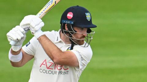 Ben Raine wearing a helmet with a Durham badge on it swooshes his bat high above his shoulder