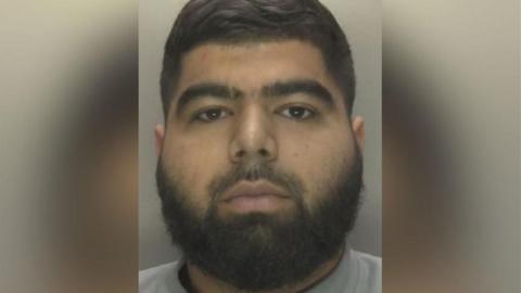 A man with black hair and a black beard against a grey background