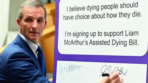 MSP Liam McArthur signs a board backing his own assisted dying bill in the Scottish Parliament. He is wearing a blue suit jacket, a blue tie and a white shirt