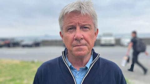 Deputy Mark Helyar - A man with grey hair looking at the camera. He is wearing a blue quarter zip jacket and a blue button up shirt.