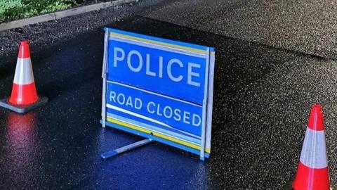 A road closed sign. The sign is blue and placed on a black tarmac road. There is an orange and white cone either side of the sign. 