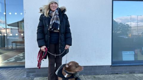 A woman in a light grey woollen hat, chequered scarf and black cold stands by a wall holding a brown bulldog on a lead.