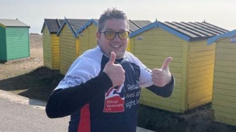 Wayne Pope wearing yellow sunglasses and a blue and white running shirt with Blind Veterans UK charity branding on it. He is running past a row of colourful beach huts and smiling at the camera, giving a double thumbs up.