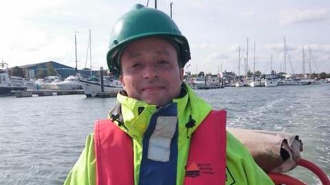 Abdulkadar Chadli in a yellow hi-vis jacket, a red lifejacket and a green hard hat. He is smiling towards the camera.