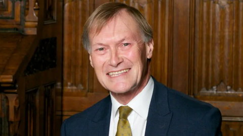 Image of Sir David Amess, smiling, wearing a navy suit and olive green tie
