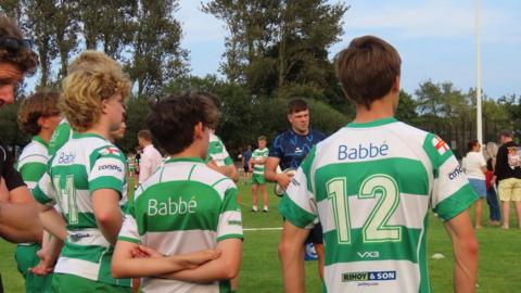 Young boys, facing away from the camera, in green and white sports shirts. There is a Bristol Bears player holding a rugby ball in front of them. The pitch is green. The sky is blue and there are trees in the background. 
