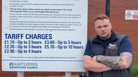 Connor Stallard is standing with his arms folded looking unhappy next to the Hartlepool Borough Council sign with new tariff changes on it. He has tattoos down one arm and is wearing a navy T-shirt under a navy gilet.