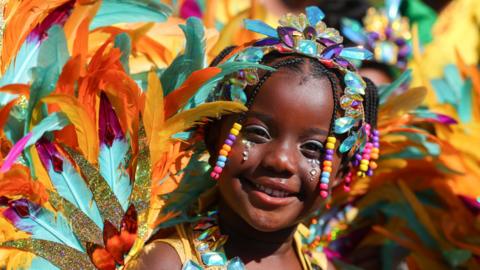 A child in brightly coloured festival attire featuring beads and feathers smiles during the Children's Day Parade.