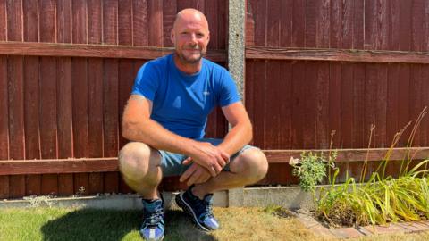 A man crouches down in his garden in front of a wooden fence. He is wearing a blue T-shirt, denim shorts and blue trainers. He has closely cropped hair and a matching beard and is smiling with lips closed.