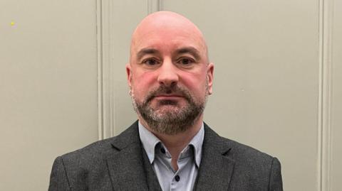 PCC Marc Jones he has a shaved head and dark beard he is wearing a charcoal three-piece suit with light grey shirt