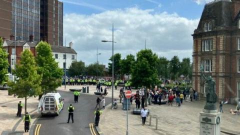 Police officers stand between two groups of people outside Hanley Town Hall in Stoke-on-Trent. There is a police van and a statue in the foreground, and a high-rise building in the background.