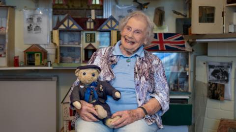 An elderly woman smiles while holding a vintage teddy bear in front of a doll's house display.