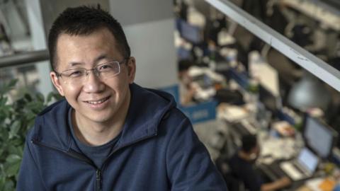 Zhang Yiming, sitting in an office. Behind him you can see people working at their desks. He is smiling, wearing glasses and a laid-back look including a t-shirt and jacket.