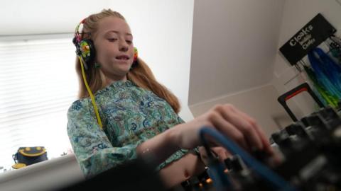 A young girl with rainbow headphones plays on DJ decks inside a house.
