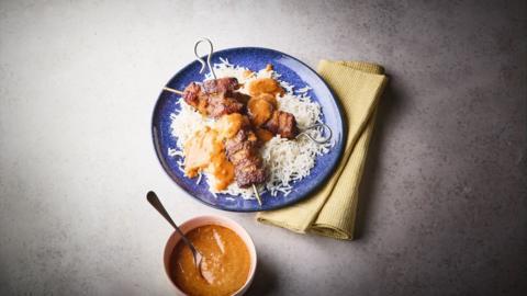 Pork skewers and rice with satay sauce on top on a blue plate. A bowl of satay sauce sits alongside.