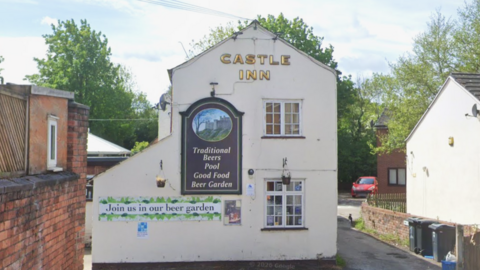 Shotton's Castle Inn - a white, two-storey pub with the name in golden letters at the top of the building. There is also a painted pub sign showing a castle on a hill with the words "traditional beers, pool, good food, beer garden" underneath. There is a redbrick wall to the left of the foreground and a house visible in the background