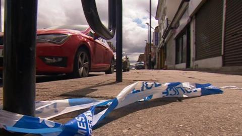 Scene of crash which shows blue and white police tape on the pavement tied on to a post with part of red car next to it on the road