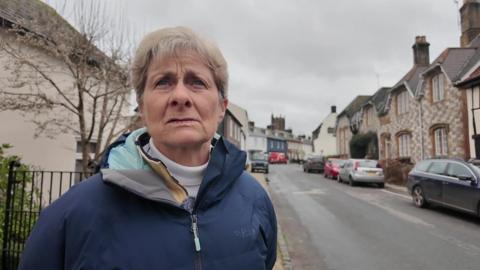 Helen Brown, a woman with short hair wearing a blue coat, stands on a pavement in the village of Cerne Abbas.