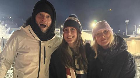 Mia Brookes standing between her mum Vicky and dad Nigel. The venue for the 2026 Winter Olympics can be seen in the background. They are all wearing head coverings and jackets.