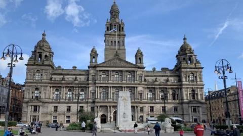 A large ornate building with a public square in front of it. The sky is blue and there are people in the square.