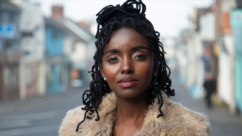 Young woman looks into the camera whilst standing on a town high street. She wears a coat with a furry collar and her black hair is half up and half down. 