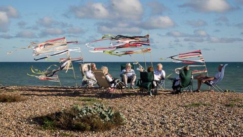 A group of retirees sitting on a pebble beach in deck chairs. There are flags and streamers next to them.