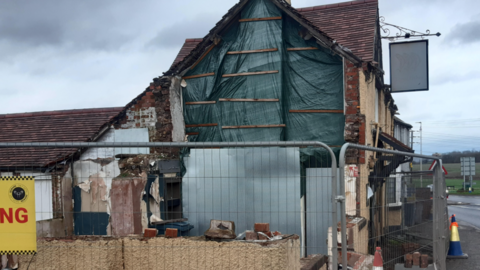 The side of a white building with tarpaulin covering one side and metal fencing surrounding it. There is a pub sign hanging from the wall