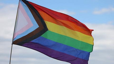 The Progress Pride Flag flying against a cloudy sky. It has the rainbow colours in stripes and a triangle at the side with a white triangle and stripes in pink, blue, brown and black