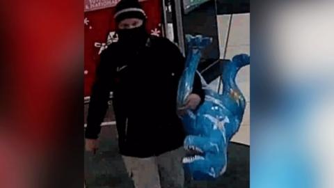 CCTV image of man in black scarf, and black hat, with only his eyes visible, and a black jacket and light grey trousers. In his left arm he is carrying a sculpture of a T-Rex, which is painted blue 