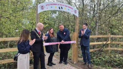 A man and a woman stand under a wooden archway in the middle of a wood, to cut a pink ribbon. Three other people next to them are clapping.