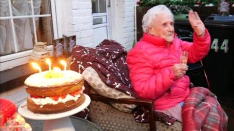 Gladys Banks, dressed in a red puffer coat with a red tartan blanket over her knees, sat in an armchair in her front garden. There is a table next to her with a huge birthday cake with four lit candles. Gladys is holding a champagne flute in one hand and smiling and waving. Out of shot is a large crowd of people stood in the street, wishing her a happy birthday