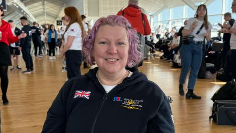 Zoe Thomas is pictured smiling in a wheelchair. She has short light purple curly hair. She is wearing a blue hoodie that has a union jack printed on the left and has the words "Invictus Games" in yellow printed on the right.